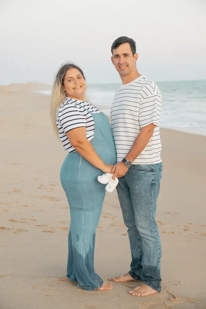 Casal grávido na praia a segurar uns sapatinhos de bebé nas mãos