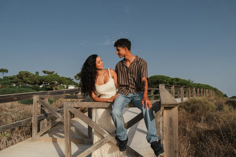 Casal sentado no corrimão de um passadiço de madeira sobre a duna, céu azul limpo