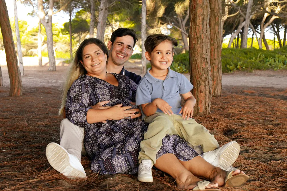 Casal com um filho pequeno sentados no chão de um pinhal, com a mãe grávida